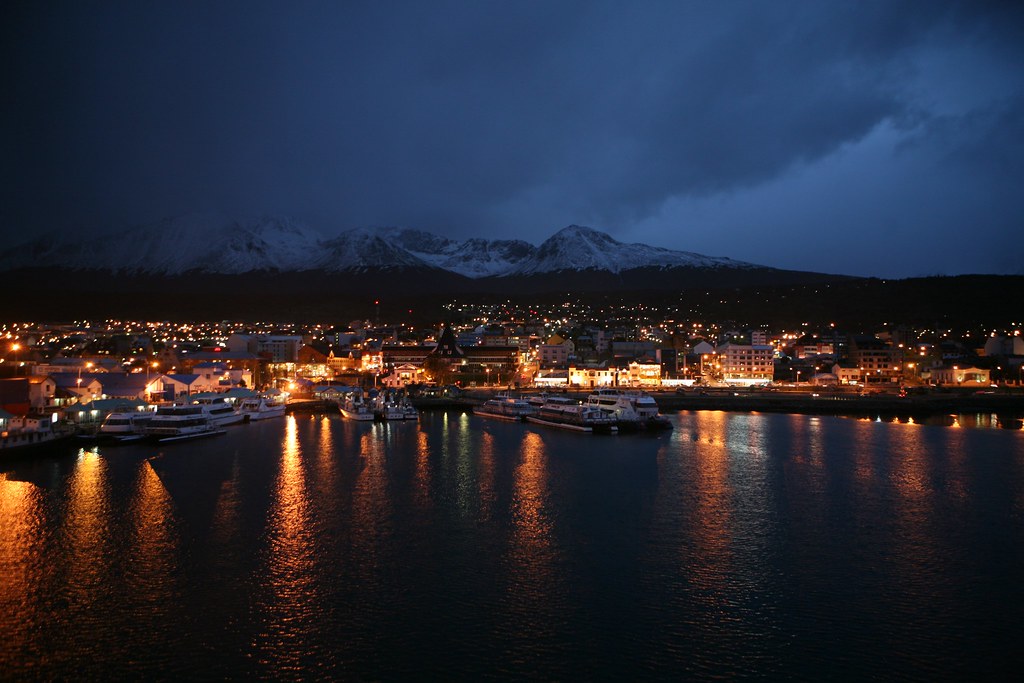 Vista noturna de Ushuaia com a cordilheira nevada ao fundo e luzes da cidade refletidas na água